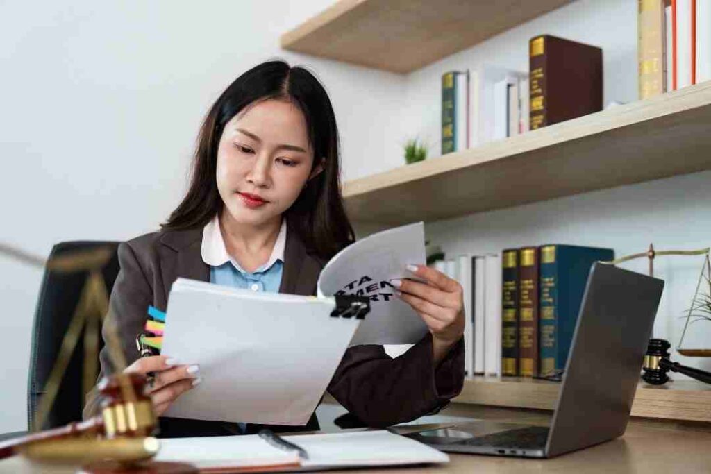 Female lawyer reading a document at a desk with a laptop and bookshelf.